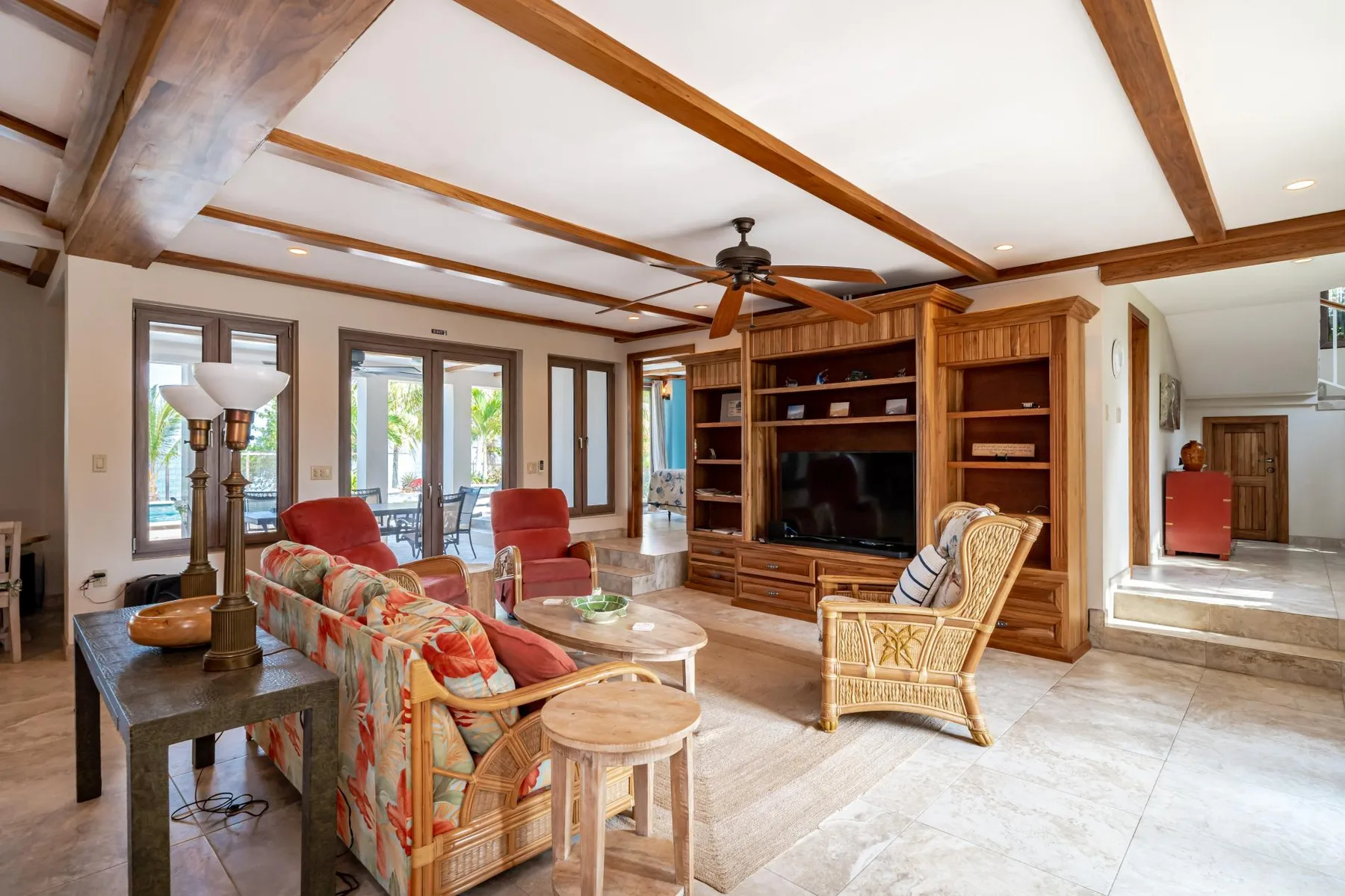A bright living room features a wooden entertainment center and seating beneath exposed ceiling beams.