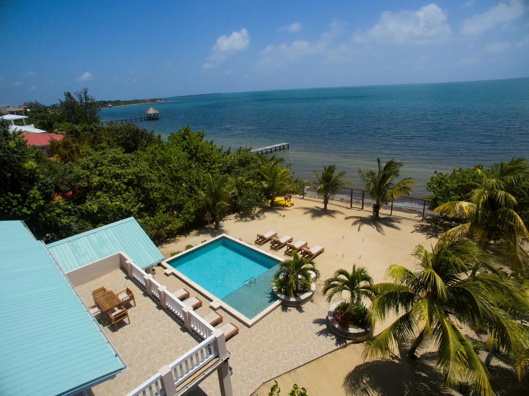 A seaside pool area overlooks a sandy beach, palm trees, and calm blue water.