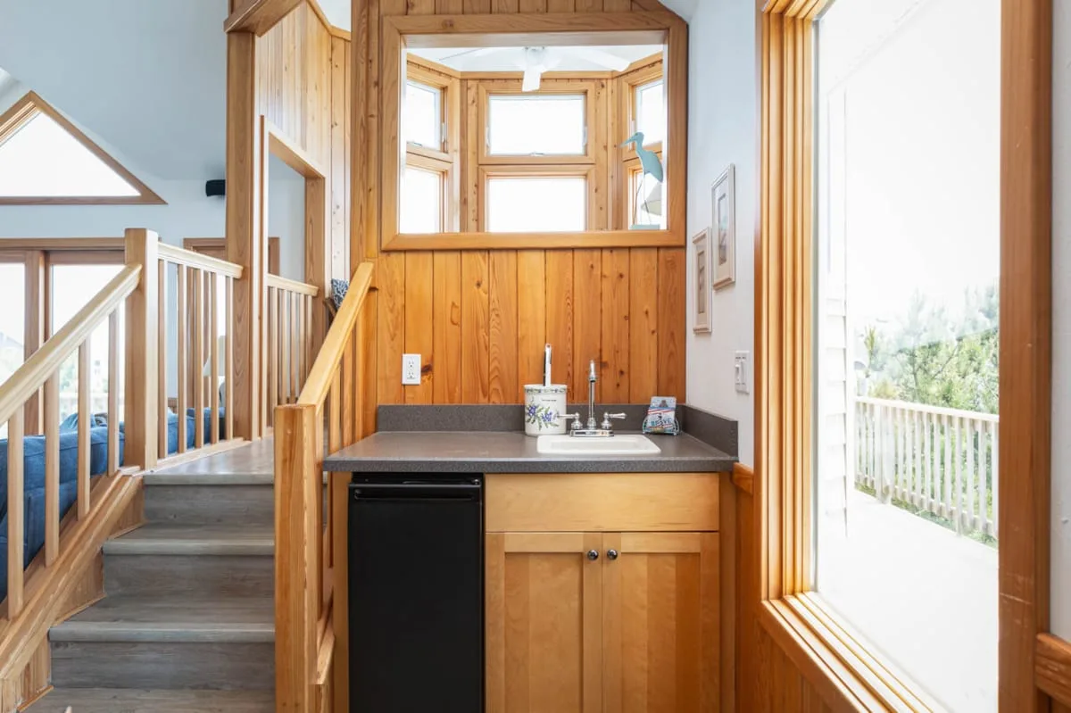 Una pequeña cocina con fregadero está junto a una escalera y una ventana luminosa.