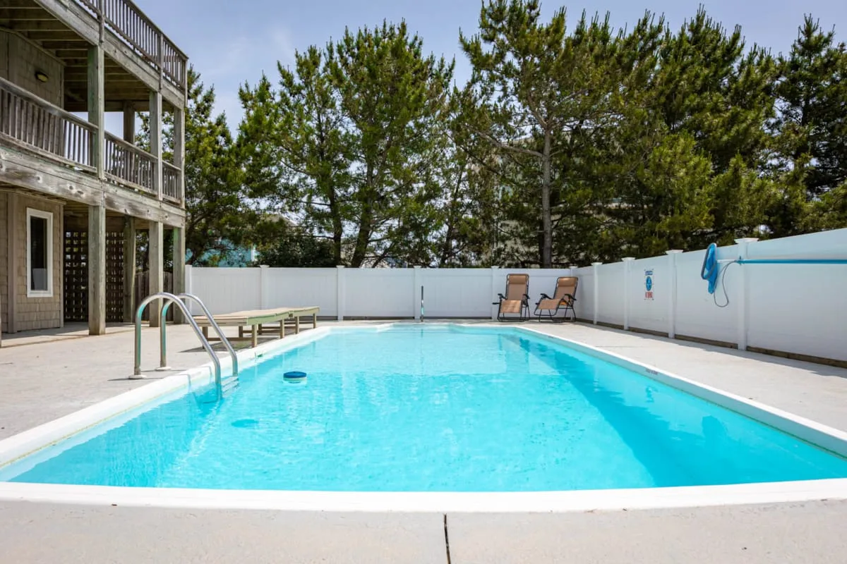 Una piscina al aire libre está rodeada por una terraza clara y una cerca blanca.