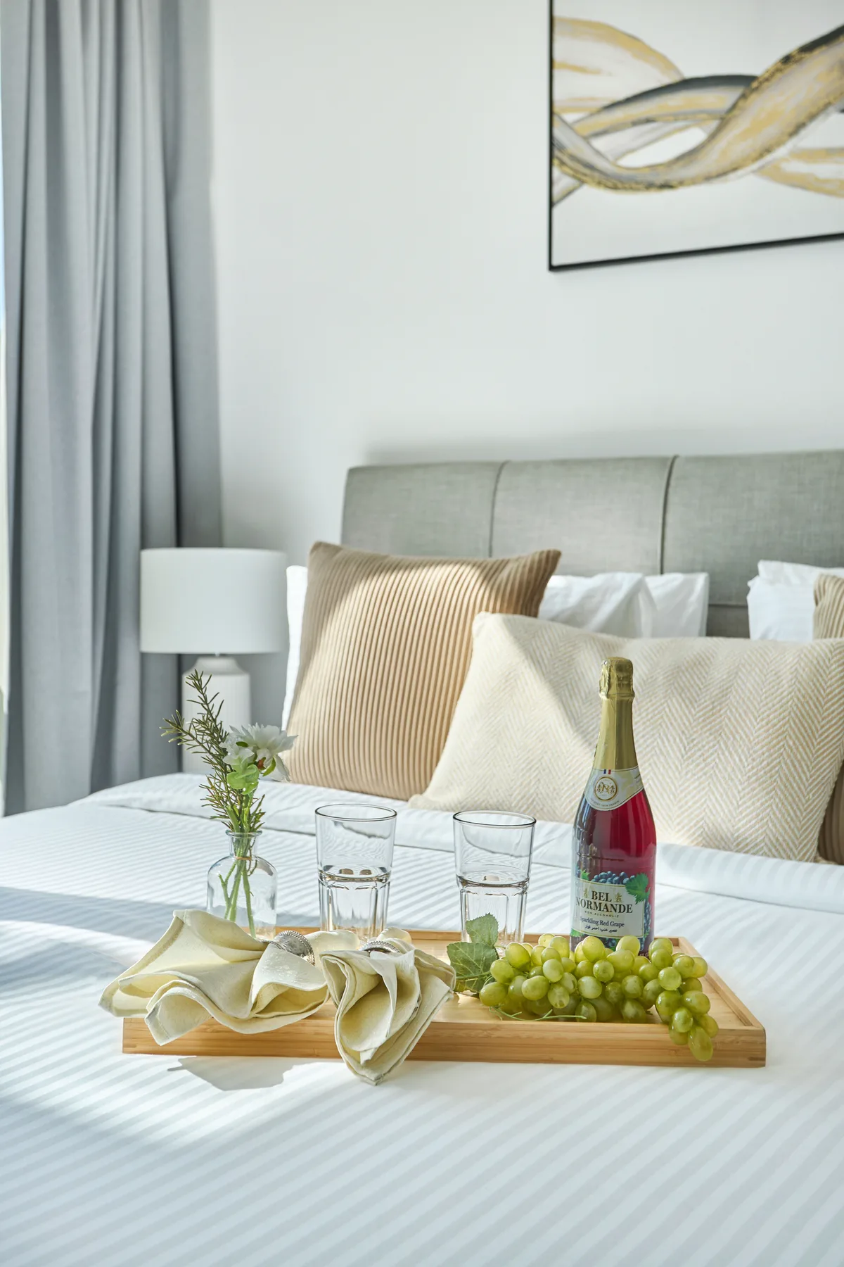 Una cama bien arreglada con cojines decorativos y una bandeja con una botella y vasos.