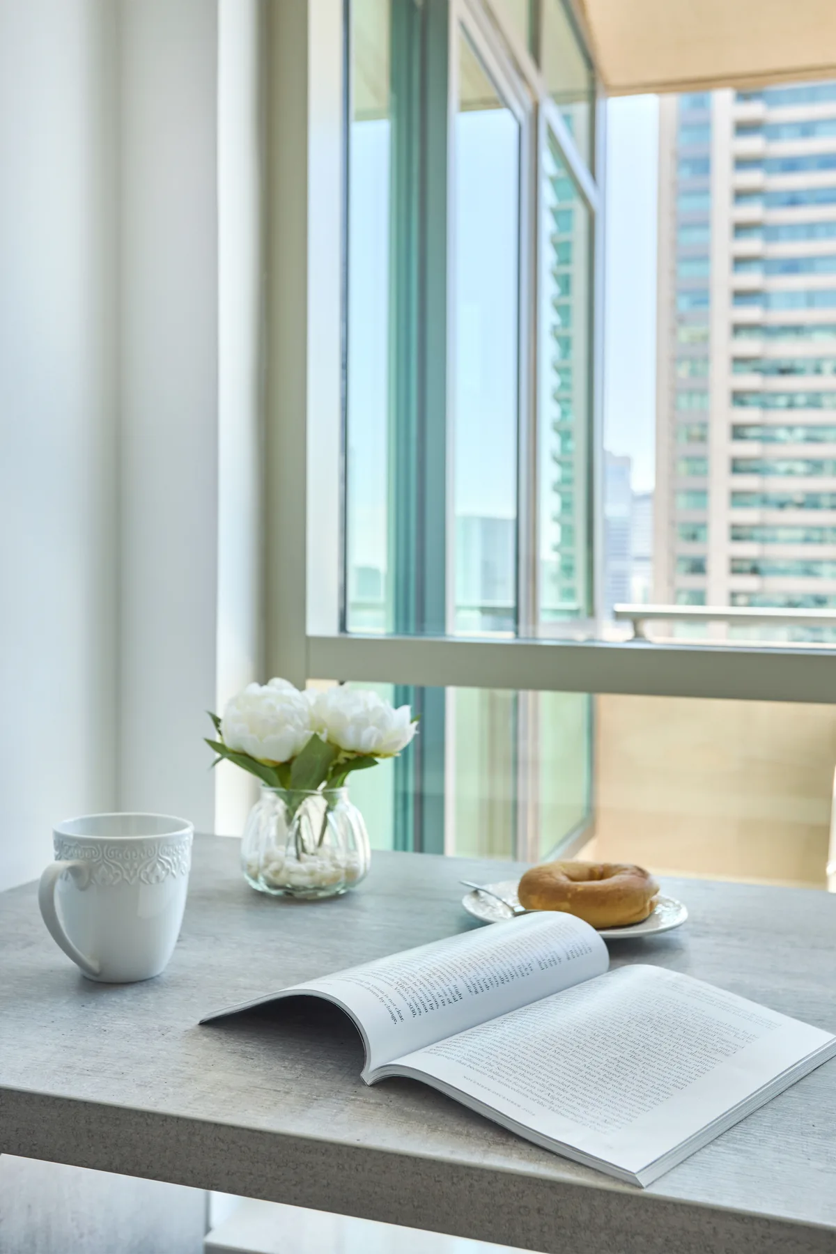 Una mesa junto a grandes ventanas sostiene un libro abierto, una taza y flores.