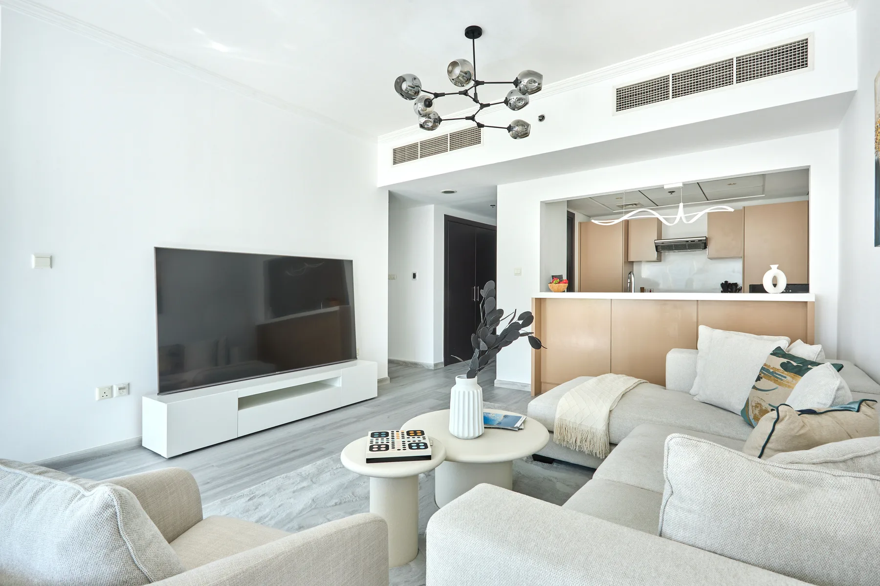 Una sala luminosa con televisor, asientos claros y una cocina abierta.