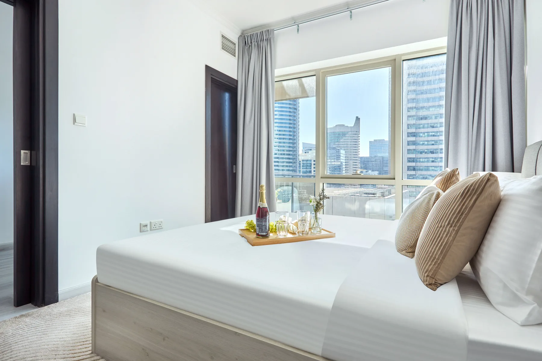 Un dormitorio luminoso con una cama tendida junto a grandes ventanas con vistas a edificios de la ciudad.