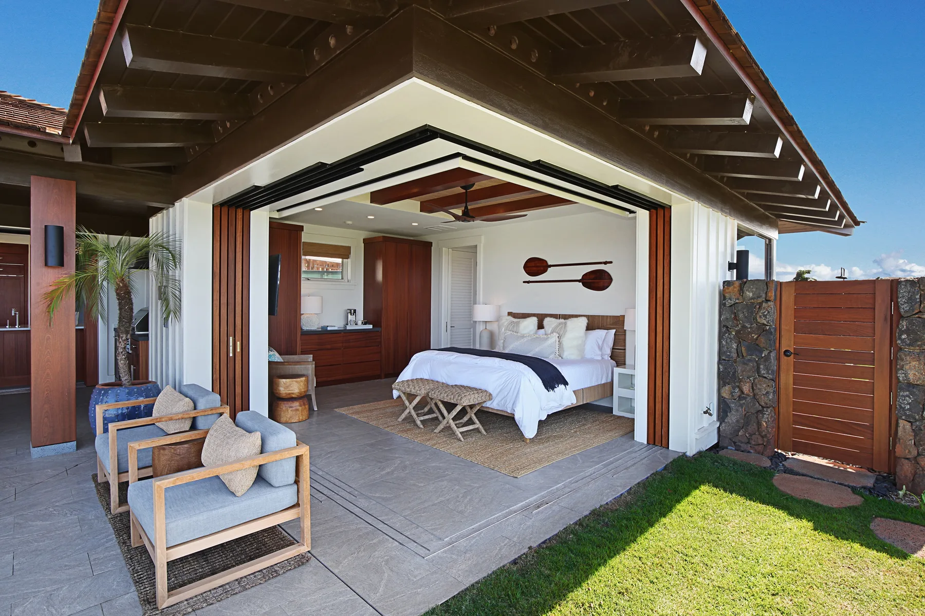An open bedroom with a bed and seating area beneath a covered patio.