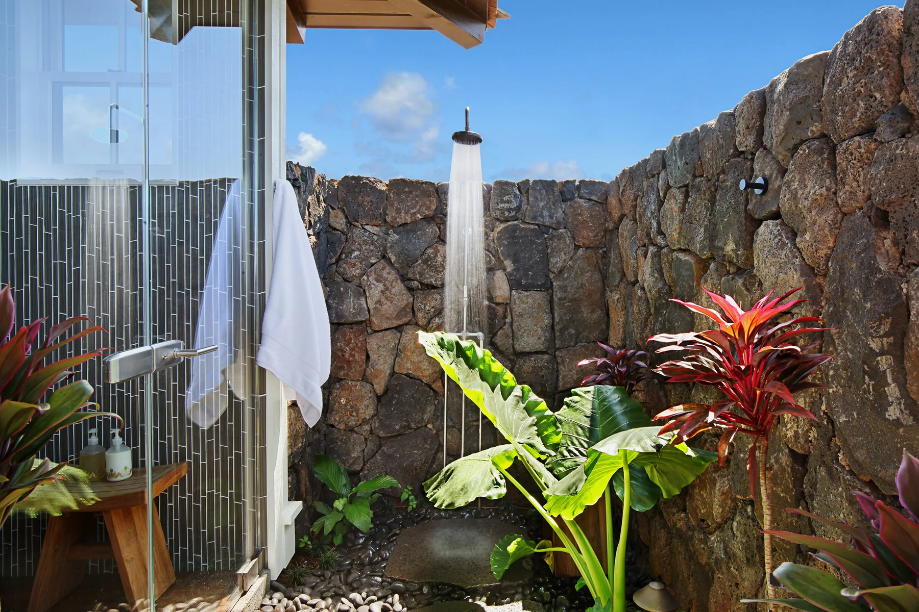 An outdoor shower area beside a stone wall, with leafy tropical plants.