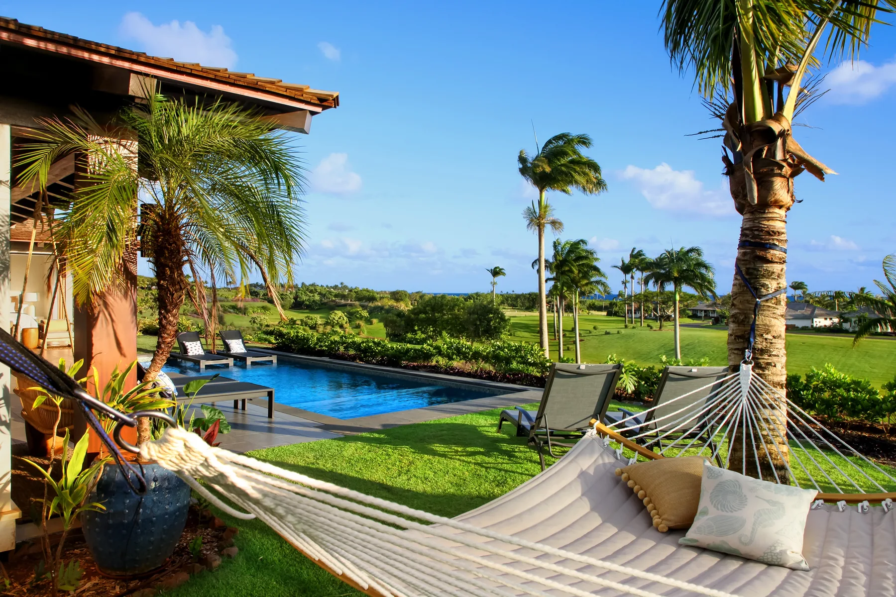 A tropical outdoor patio overlooks a pool and green lawn, with a hammock in the foreground.