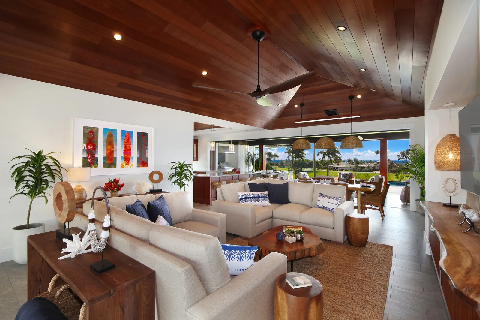 An open-plan living room has cushioned seating, a wooden ceiling, and windows overlooking greenery.