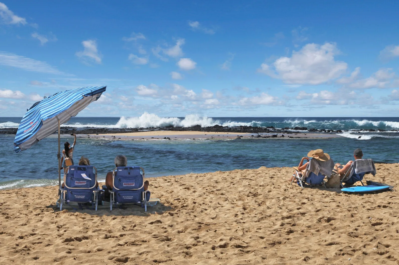 A sandy beach overlooks ocean waves, with beachgoers seated under a striped umbrella.