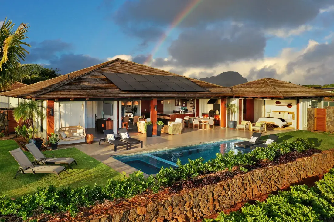 A single-story house opens onto a swimming pool and outdoor seating beneath a rainbow.
