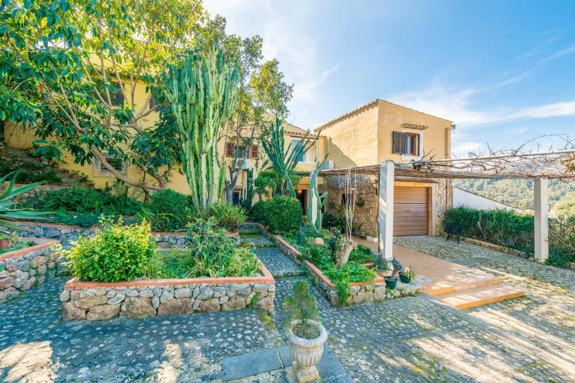 A tan house stands beside a stone-paved driveway and landscaped garden.