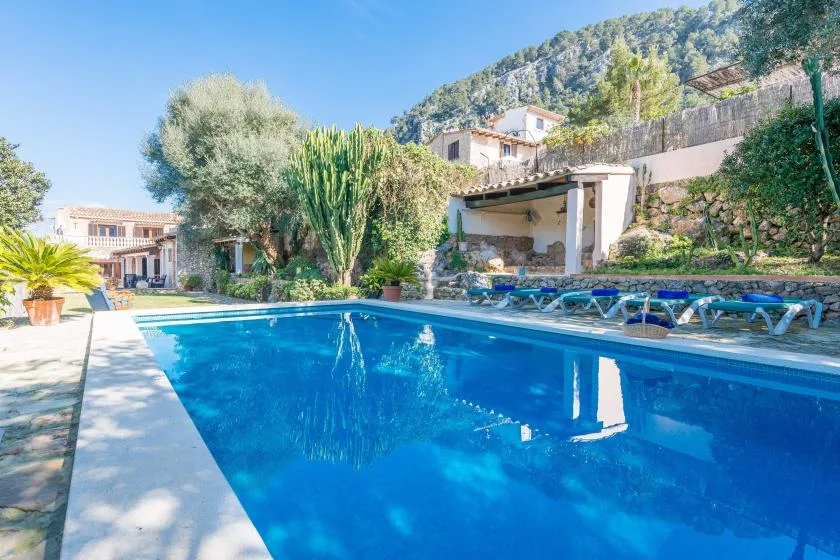 A swimming pool sits beside a landscaped terrace with loungers and hillside buildings.