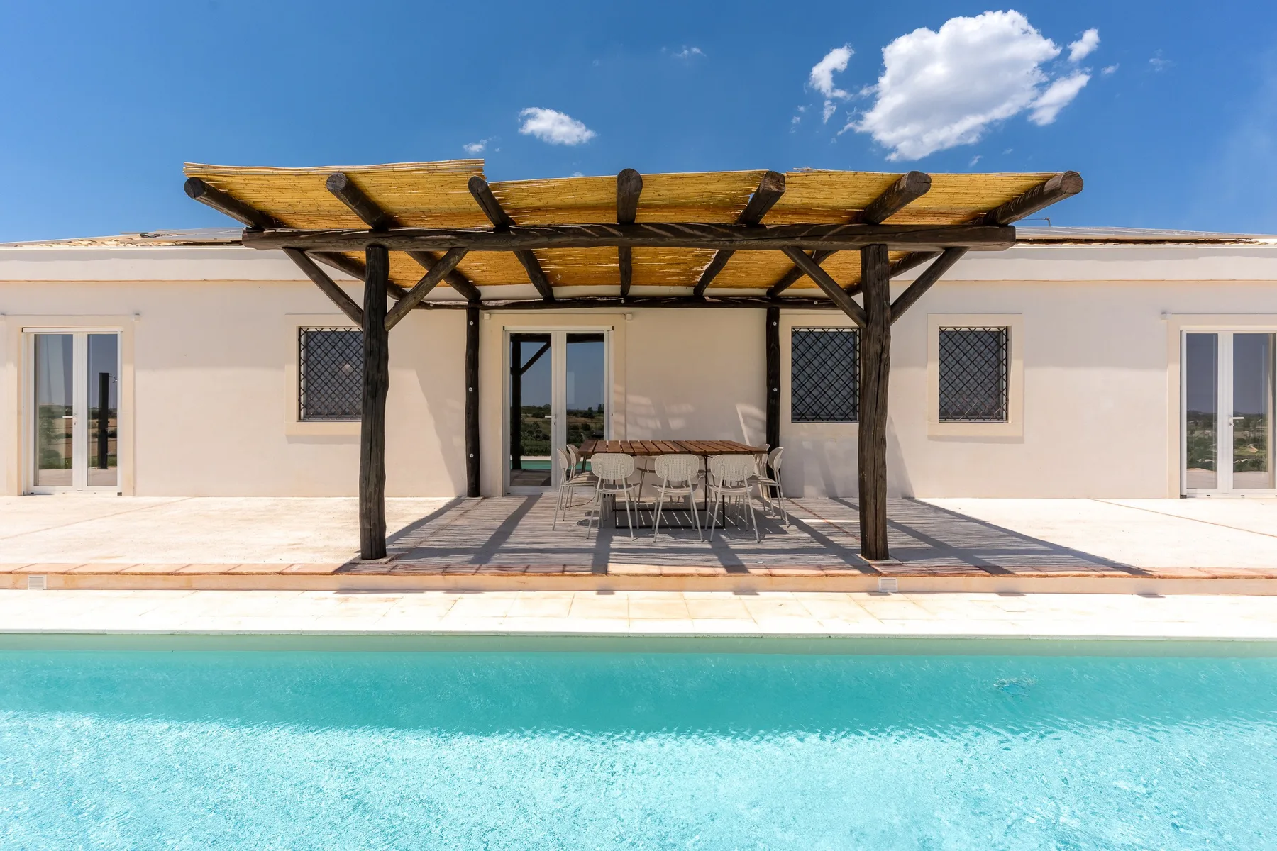 Una piscina da a un patio con asientos al aire libre bajo una pérgola de madera.