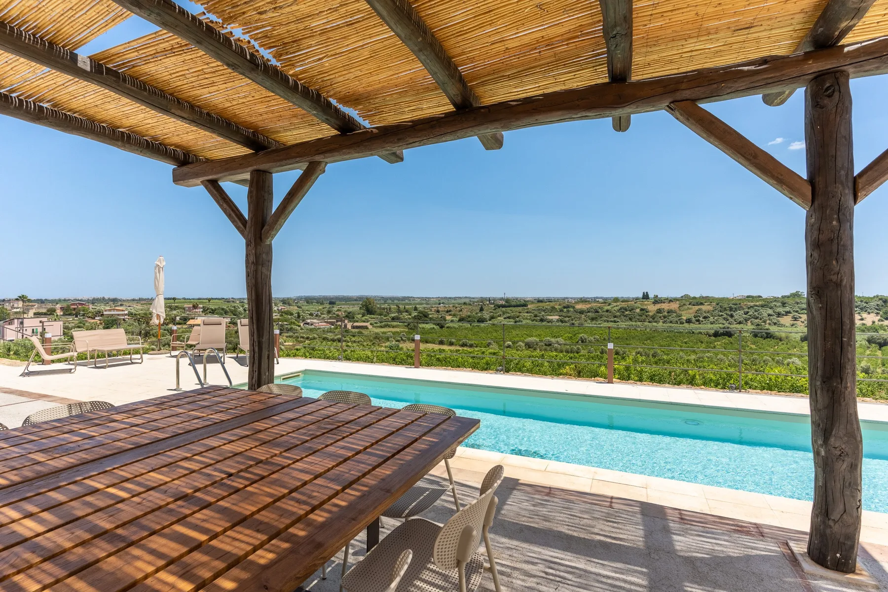 Un patio a la sombra con una mesa da a una piscina y al campo verde.