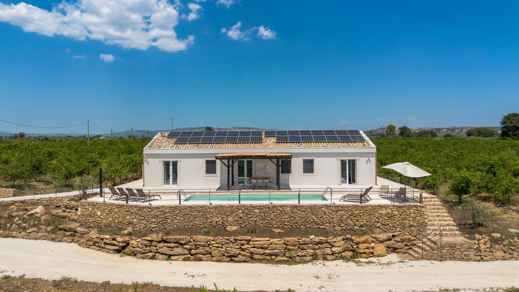 Un edificio blanco bajo con paneles solares se alza tras una piscina y una terraza de piedra.