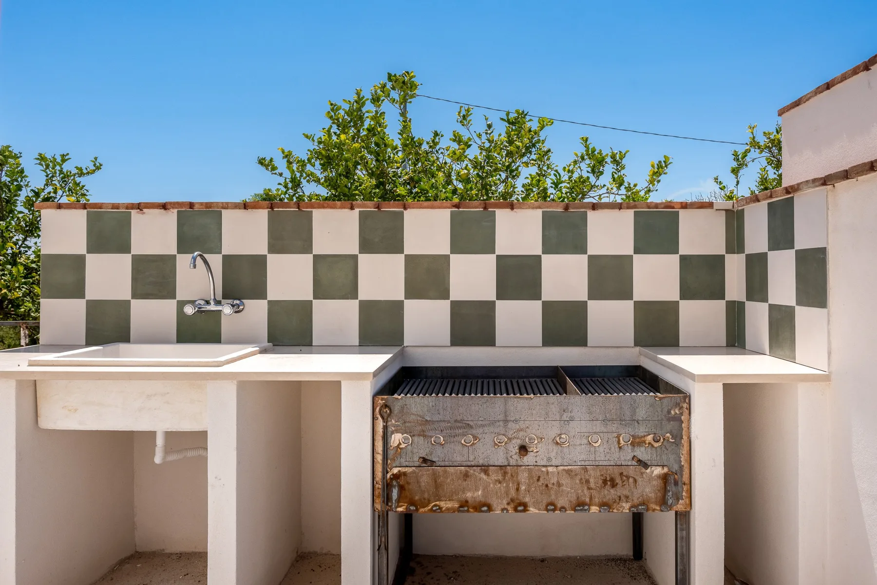 Una zona de cocina al aire libre con parrilla y fregadero bajo un salpicadero de azulejos a cuadros.