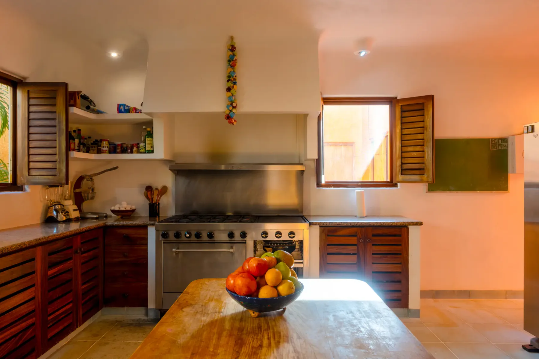Cocina con iluminación cálida, una cocina grande, estantes abiertos, dos ventanas de madera y un frutero sobre una mesa de madera en primer plano.