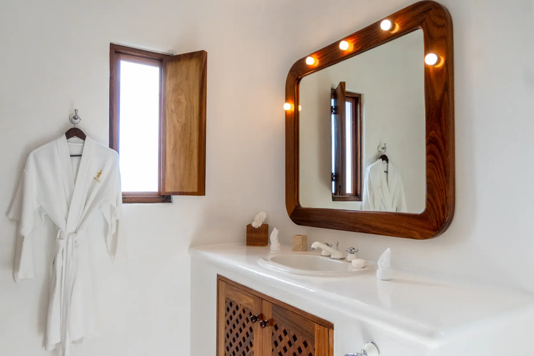 Tocador de baño con espejo iluminado, lavabo blanco y ventana de madera abierta.
