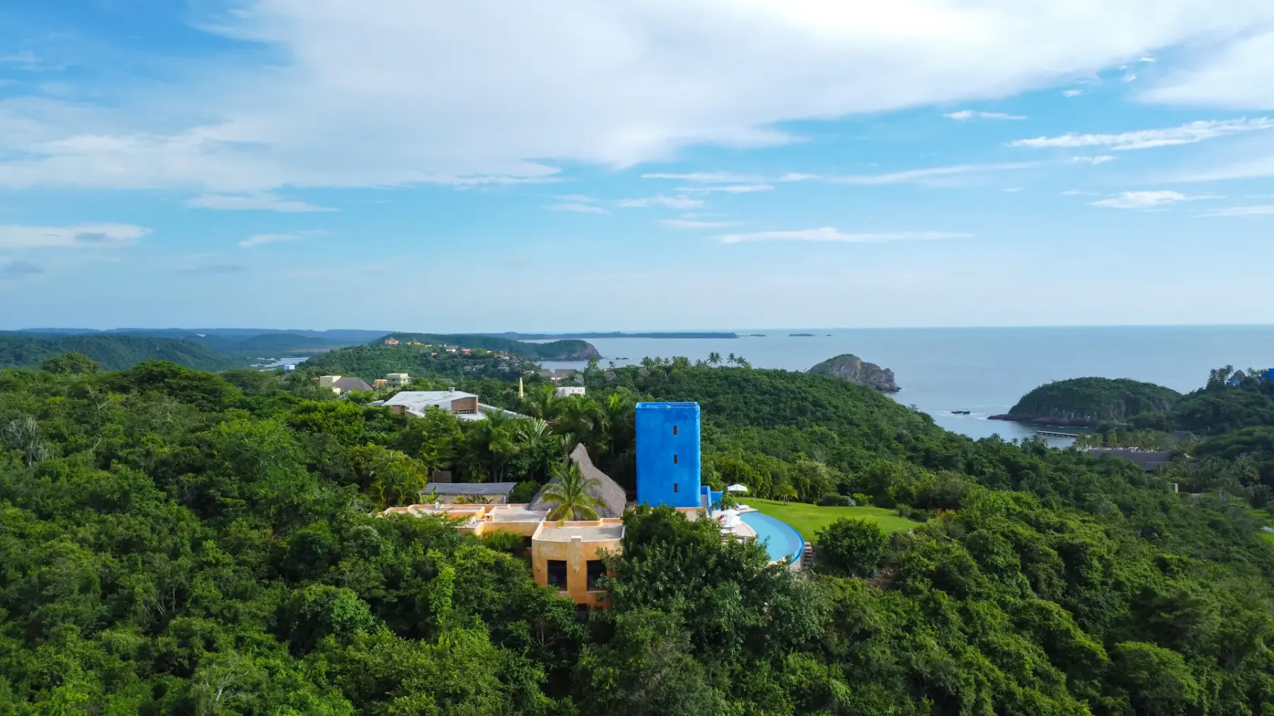 Vista aérea de una torre azul entre frondosos árboles verdes, con la costa y el océano al fondo bajo un cielo azul con nubes.