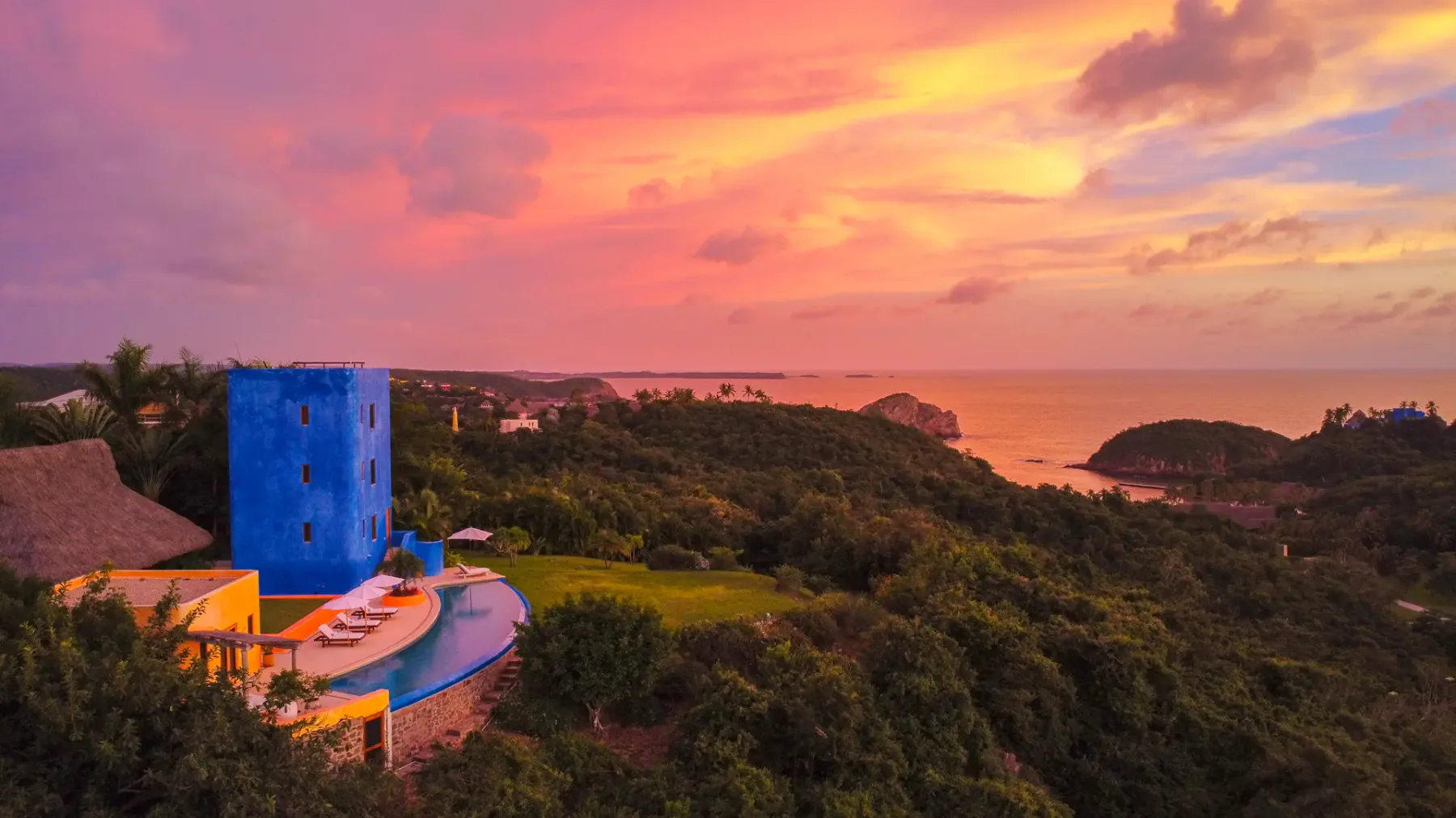 Un edificio azul y una piscina curva dominan un paisaje costero verde bajo un cielo colorido al atardecer.