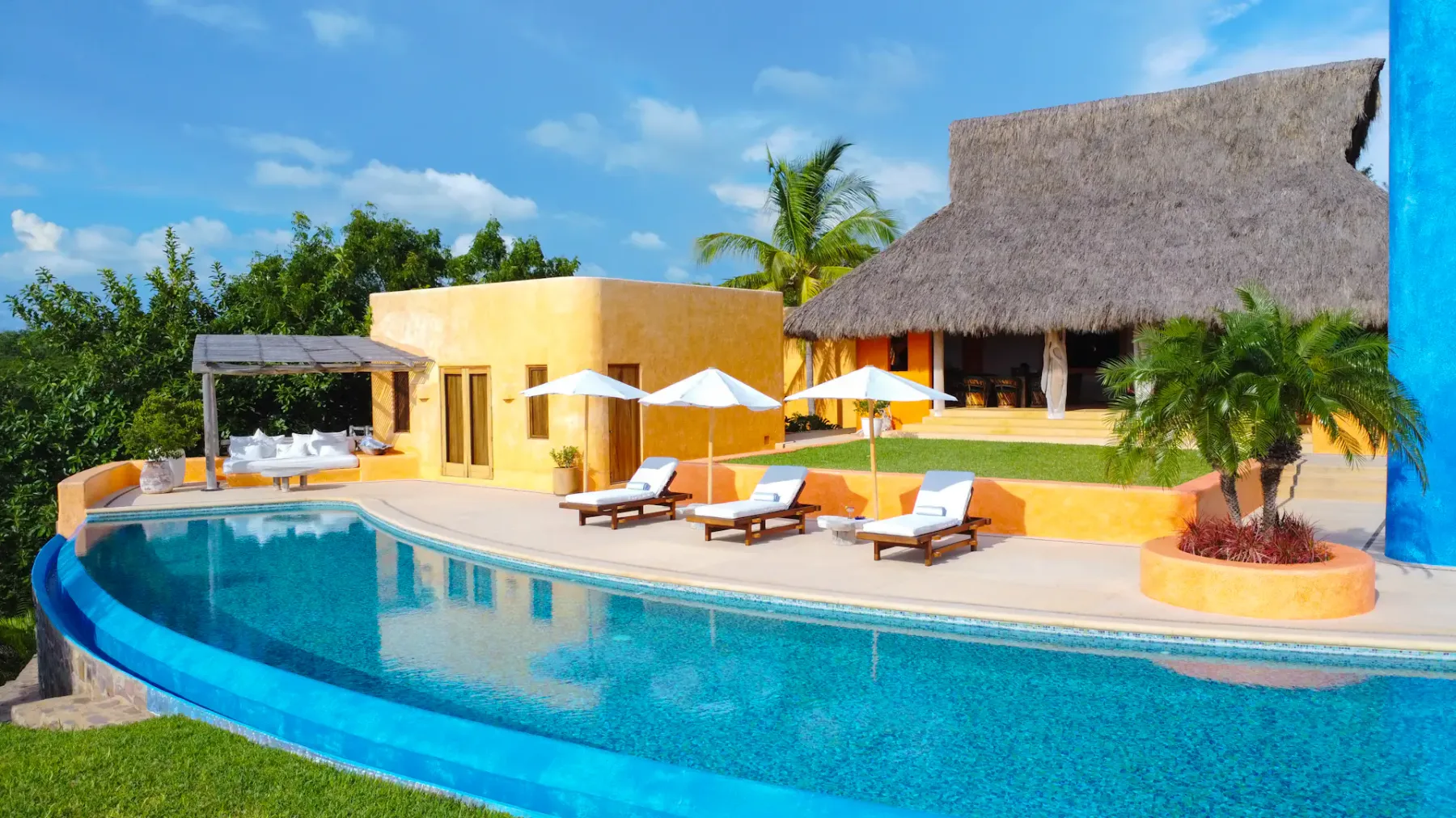 Villa tropical con techo de paja junto a una piscina curva, rodeada de palmeras y tumbonas bajo sombrillas blancas en un día soleado.