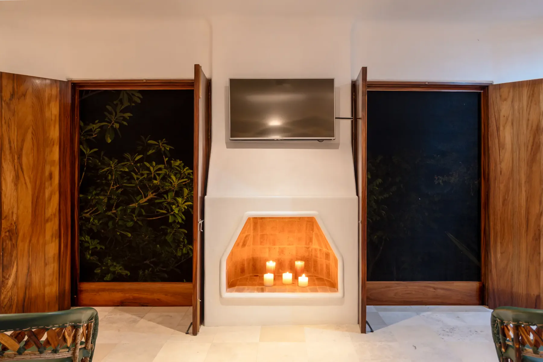 Interior moderno con un televisor montado en la pared sobre una chimenea empotrada con velas, flanqueada por puertas de madera con vistas a las ramas de los árboles del exterior.