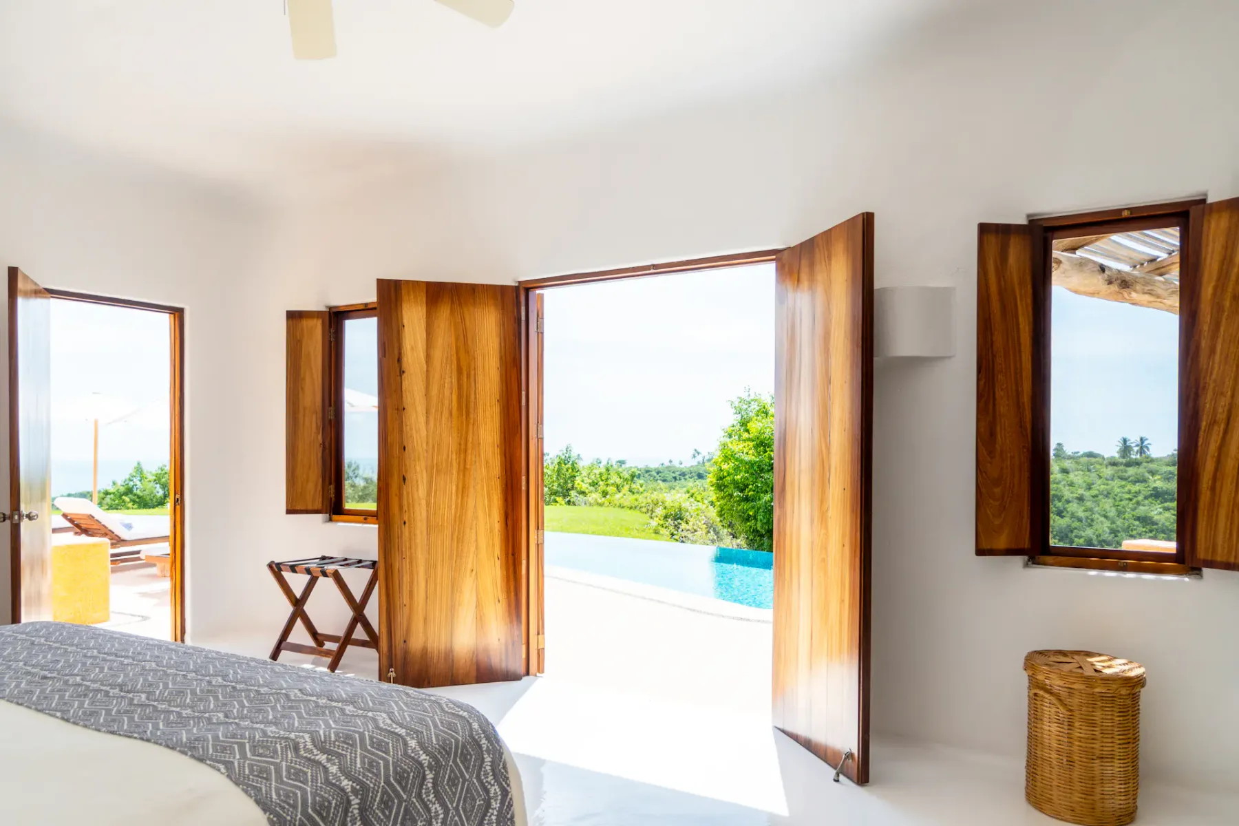 Dormitorio luminoso con puertas de madera abiertas que dan a un patio con vegetación y una piscina. La luz natural llena la habitación de paredes blancas.