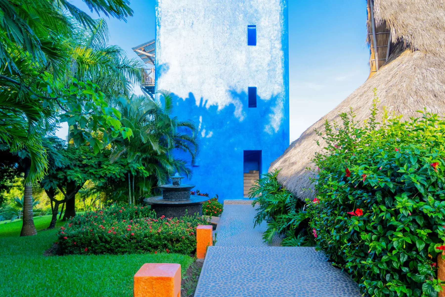 Edificio de azul intenso rodeado de vegetación exuberante, un sendero de piedra y plantas tropicales bajo un cielo azul despejado.