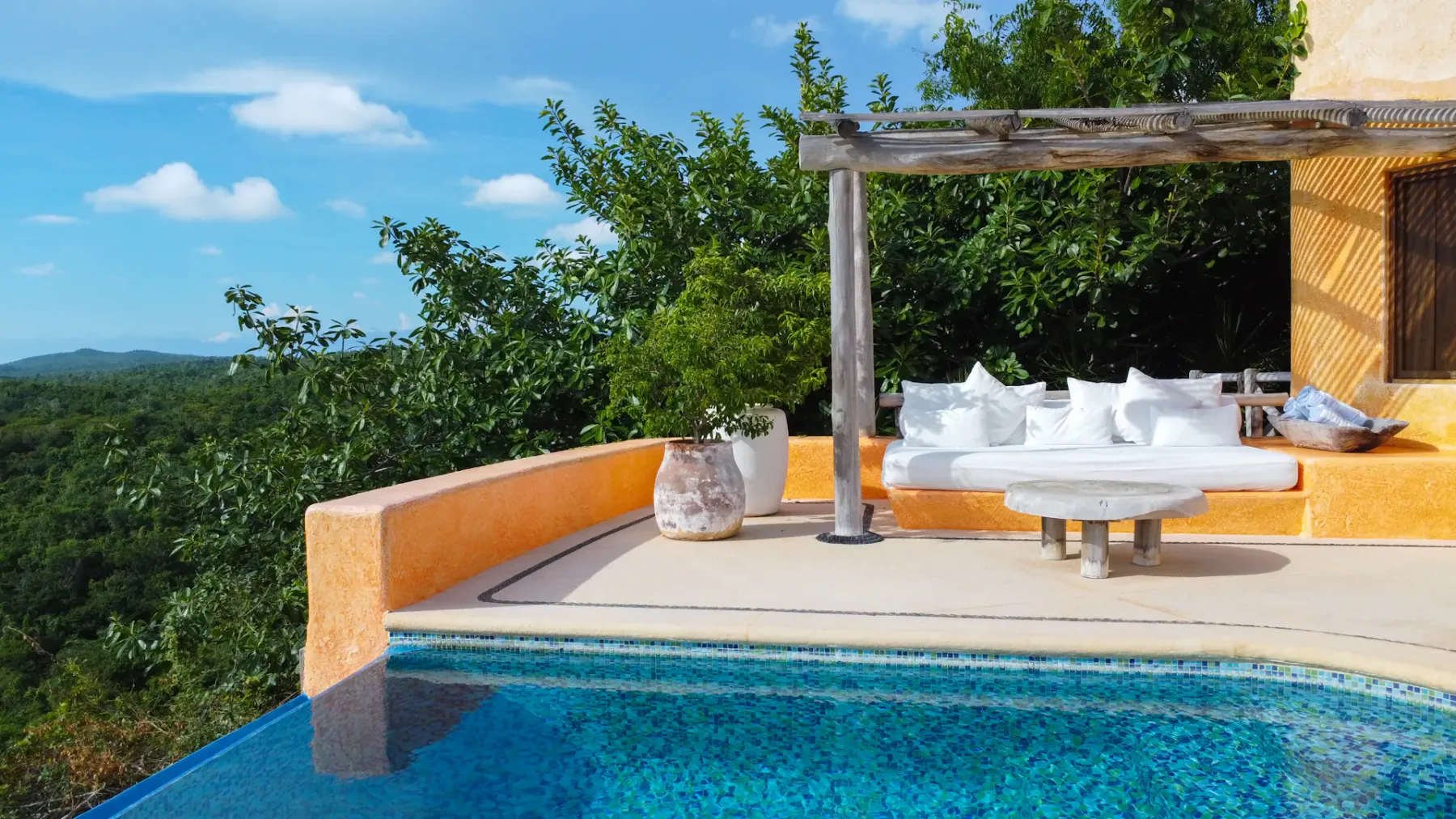 Patio junto a la piscina con asientos blancos acolchados bajo una pérgola de madera, con vistas a colinas verdes.