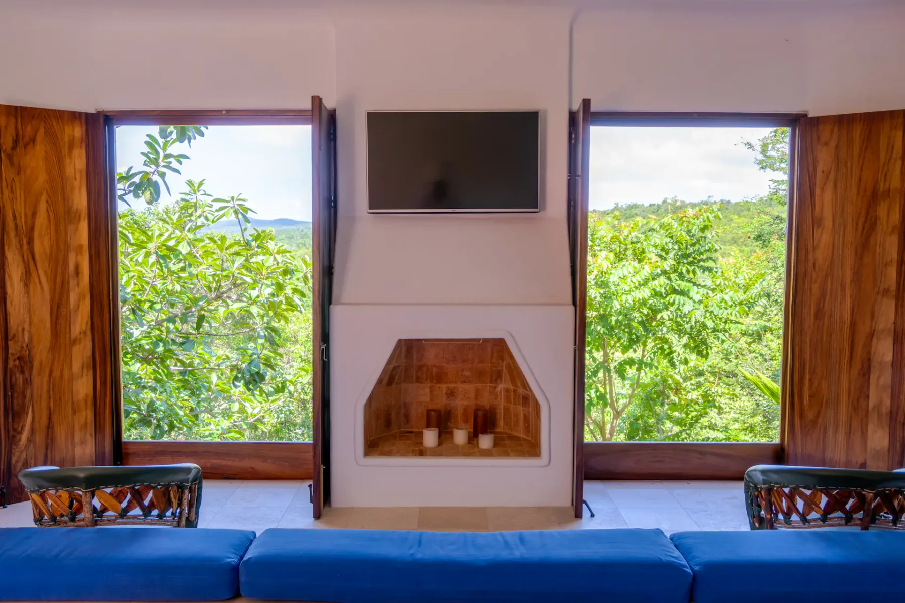 Sala de estar con un televisor montado en la pared, una chimenea y ventanas con vistas a la vegetación.