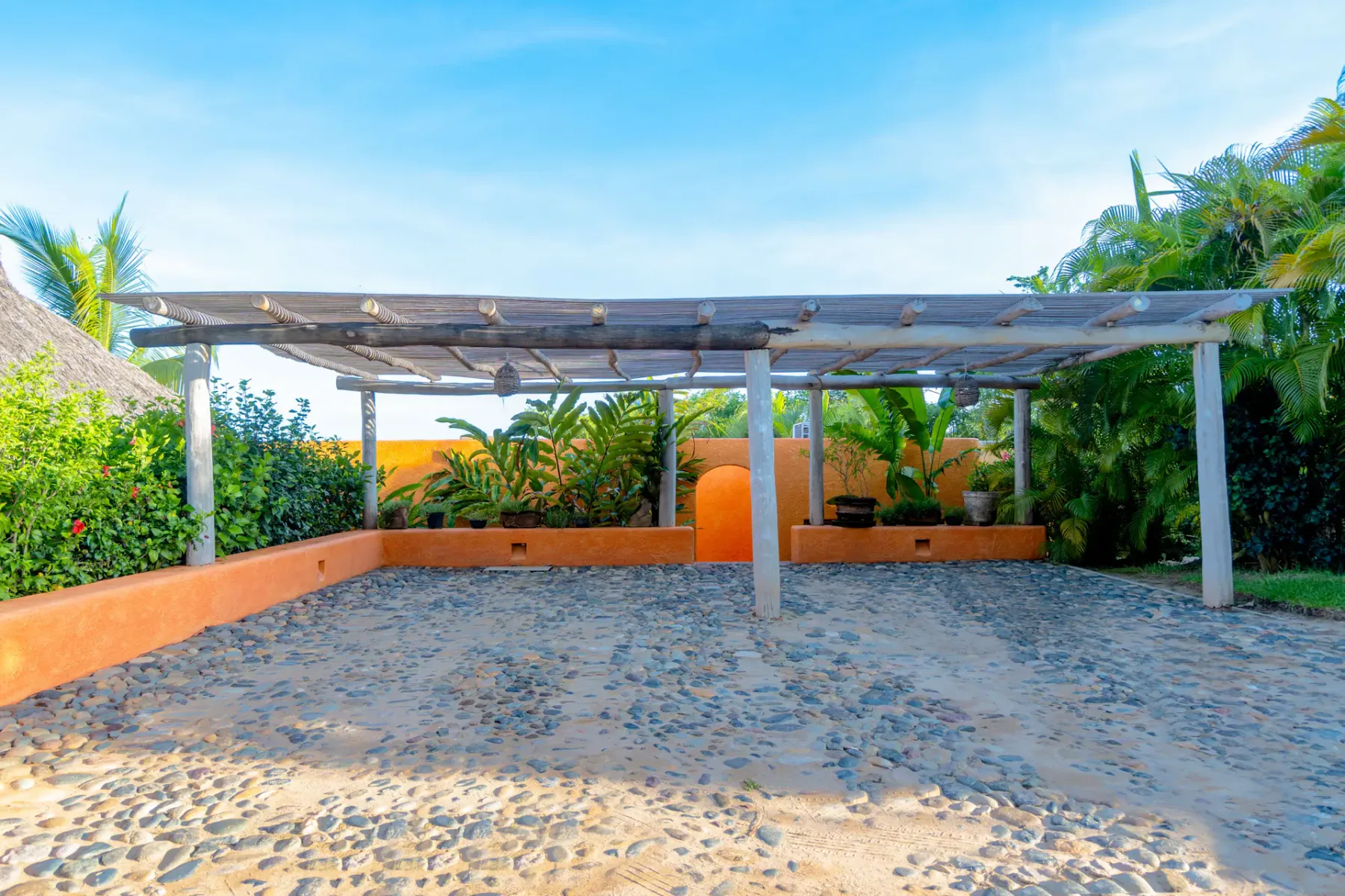 Una pérgola con techo de madera se alza sobre un patio de piedra, rodeada de exuberantes plantas verdes y paredes naranjas bajo un cielo azul despejado.
