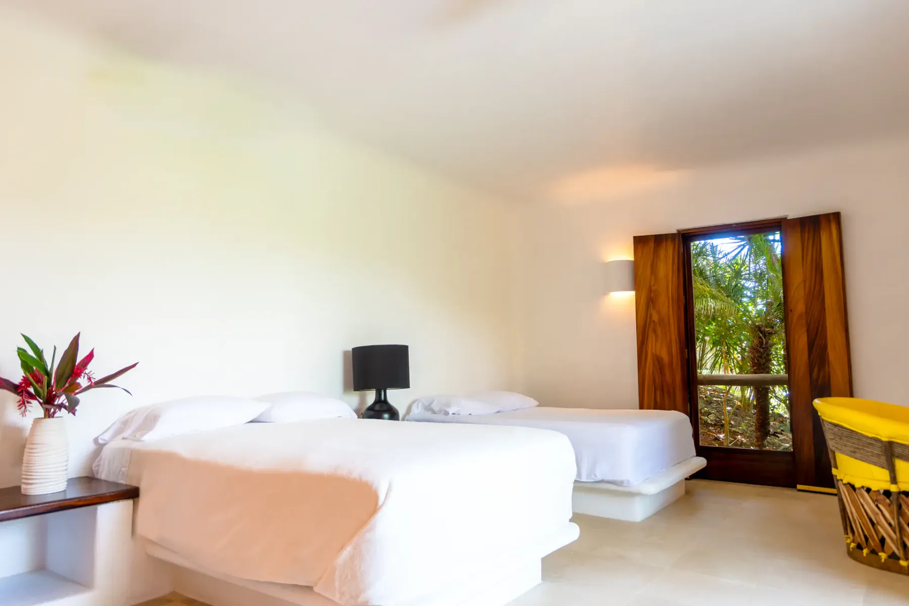 Dormitorio minimalista con dos camas, una lámpara negra y un jarrón con flores rojas. Una ventana ofrece vistas a la exuberante vegetación exterior.
