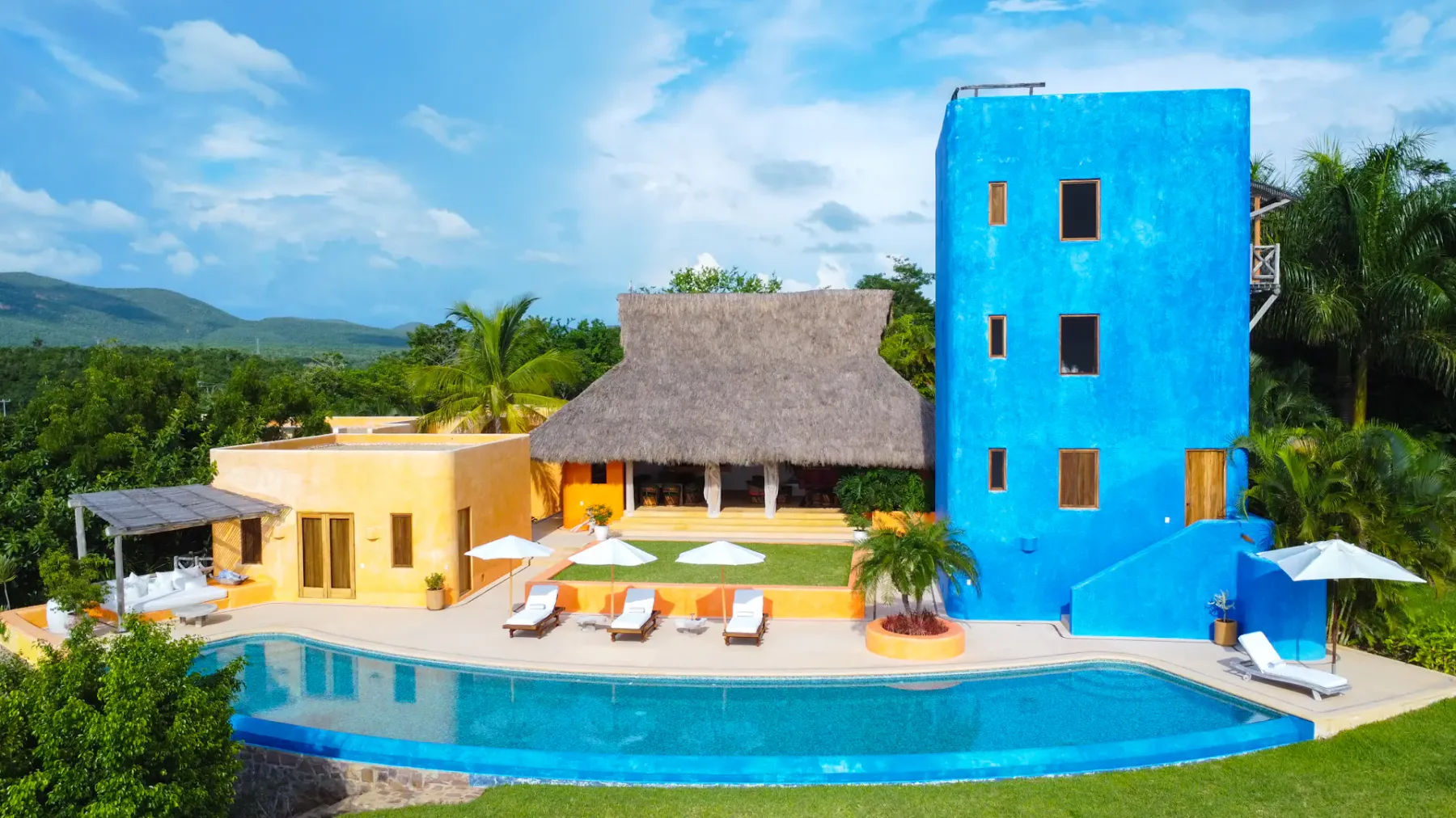 Villa colorida con techo de paja, una torre azul, un edificio amarillo, piscina y tumbonas en un entorno tropical exuberante.