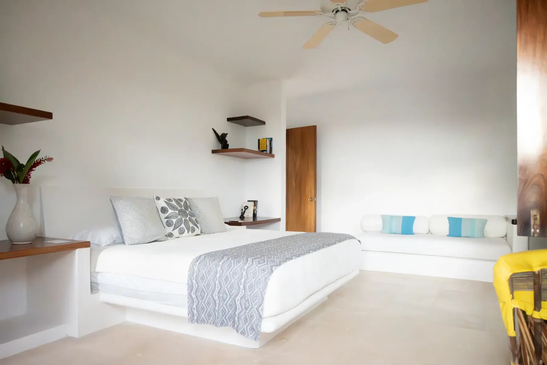 Un dormitorio luminoso con una cama blanca, una manta gris, un ventilador de techo y estantes.