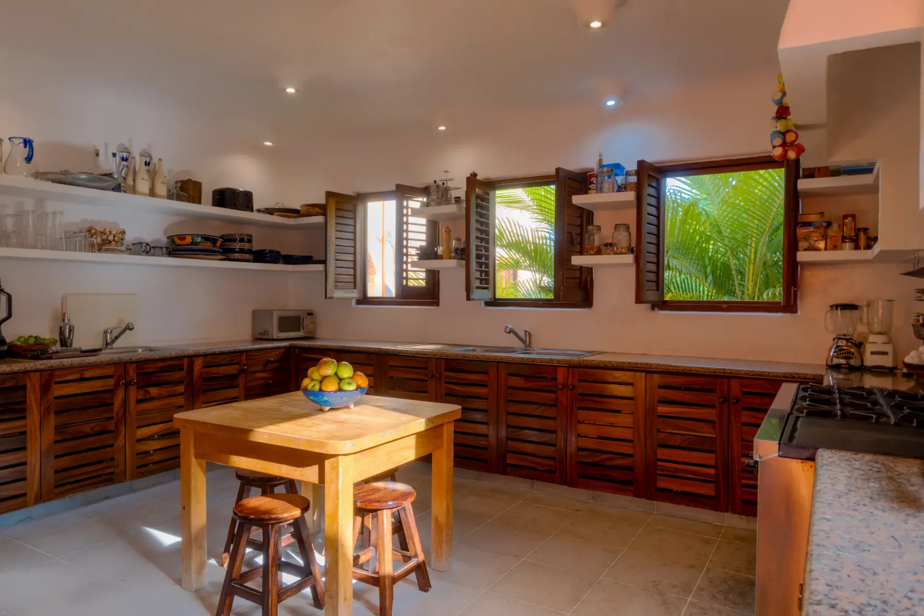 Cocina soleada con armarios de madera, estantes abiertos, una mesa central con taburetes y ventanas con vistas a la exuberante vegetación exterior.