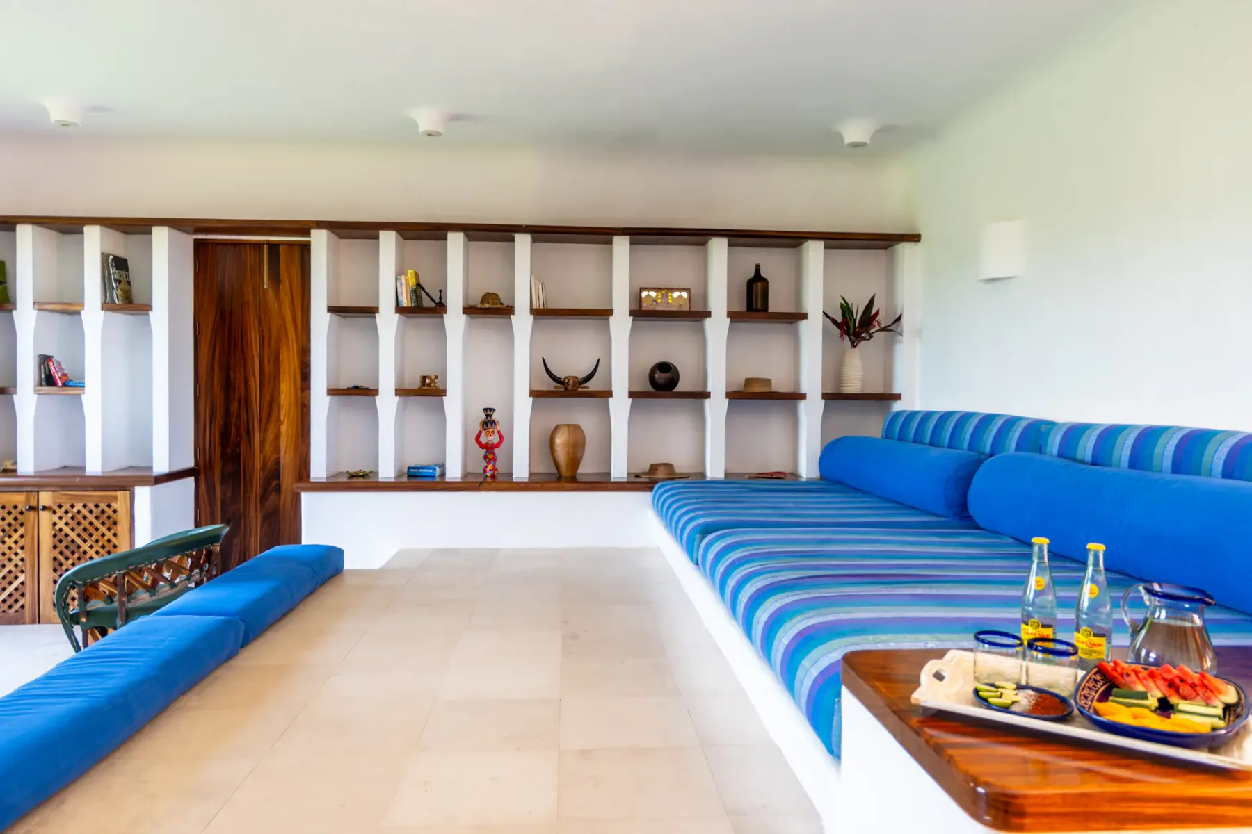 Sala con sofás de rayas azules, paredes blancas, estantes de madera y una mesa pequeña con aperitivos y bebidas. Decoración minimalista.