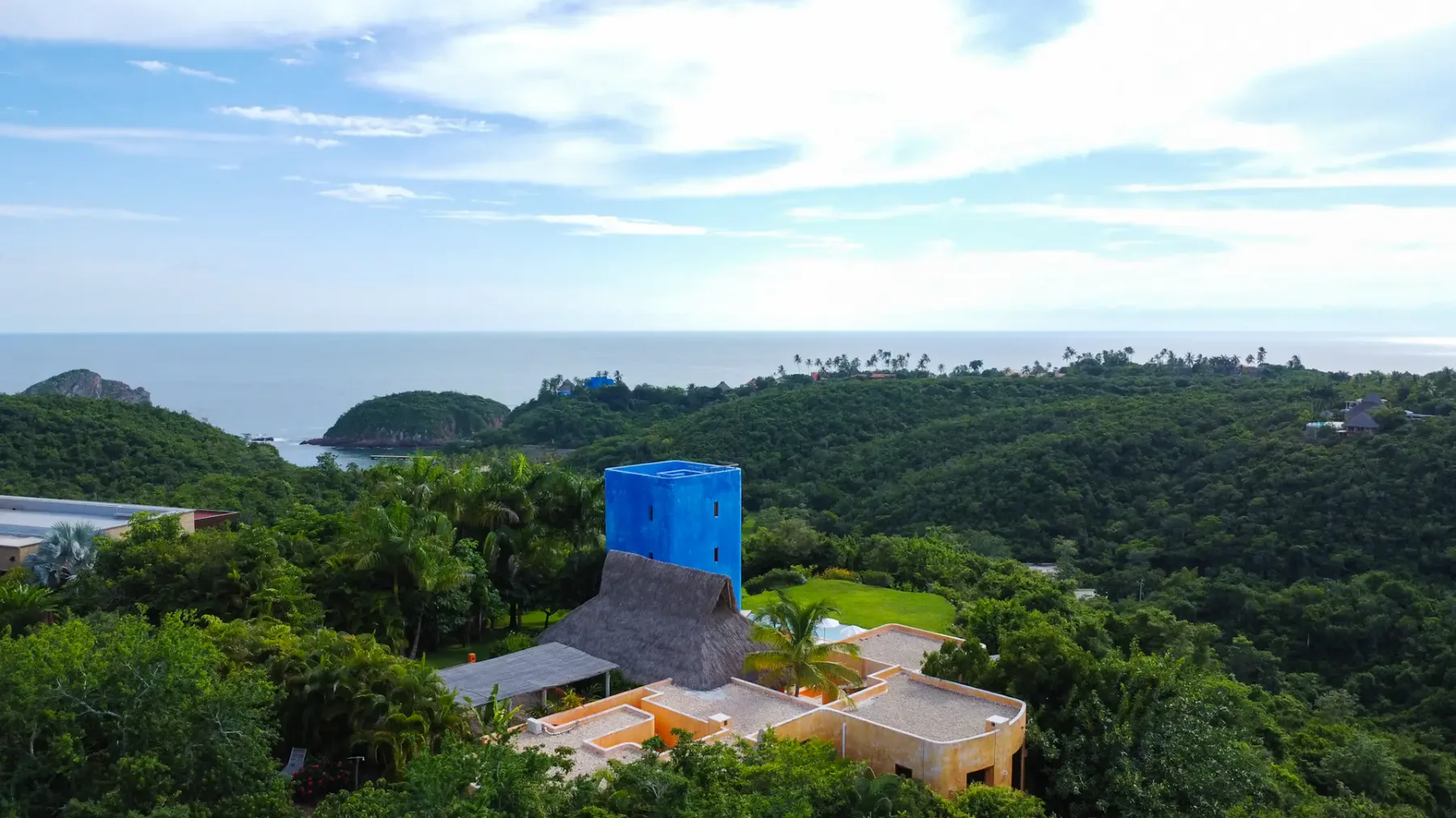 Una torre de azul intenso se eleva sobre un paisaje de exuberante vegetación cerca del océano, rodeada de otros edificios coloridos y denso follaje.