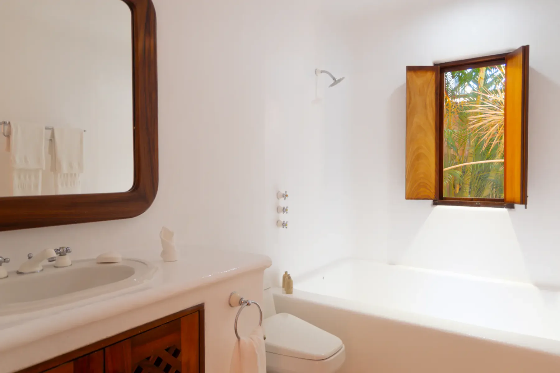 Baño minimalista con paredes blancas, un espejo con marco de madera, una bañera, una ventana pequeña con contraventanas de madera abiertas y exuberante vegetación en el exterior.