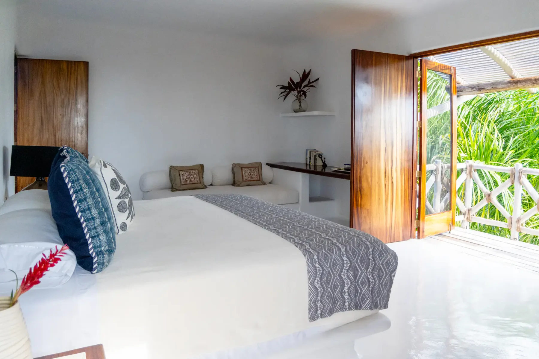 Dormitorio luminoso con una cama grande, cojines de colores y puertas de madera que dan a un balcón con vistas a una vegetación exuberante.