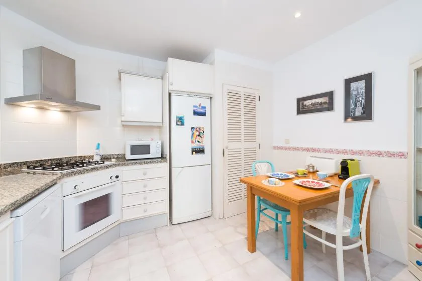 Una cocina luminosa con armarios blancos, electrodomésticos y una mesa de comedor de madera.