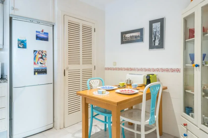 Un comedor luminoso tiene una mesa de madera y sillas de colores junto a un refrigerador.