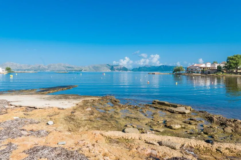 Una orilla rocosa bordea aguas azules tranquilas, con colinas lejanas bajo un cielo despejado.
