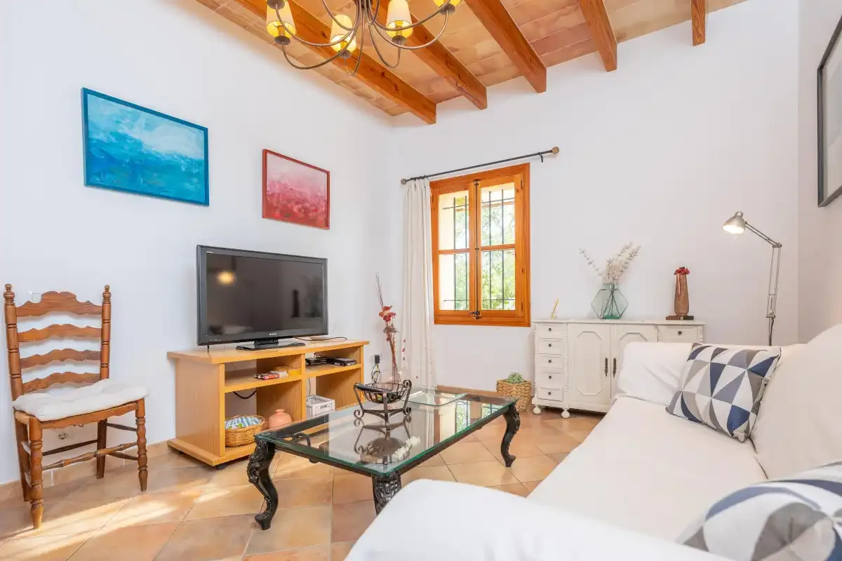 Una sala de estar luminosa con asientos blancos, un televisor y vigas de madera vistas en el techo.