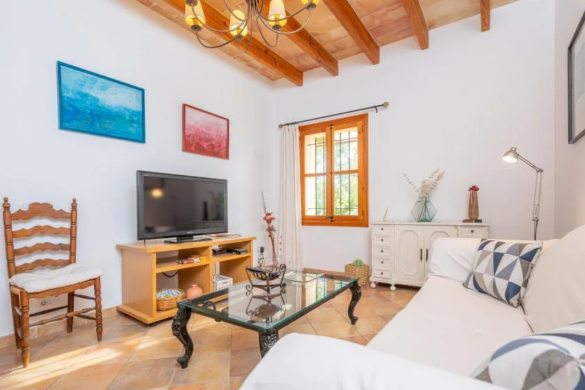 Una sala luminosa tiene un televisor, una mesa de centro de vidrio y asientos blancos.