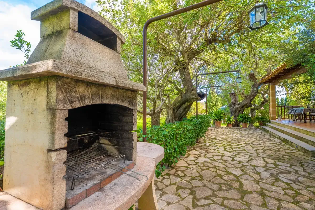 Un patio exterior pavimentado con piedra, una chimenea empotrada y árboles frondosos.