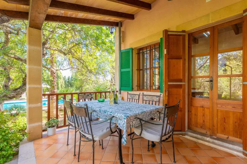 Un patio cubierto tiene una mesa de comedor y sillas junto a puertas de madera abiertas.