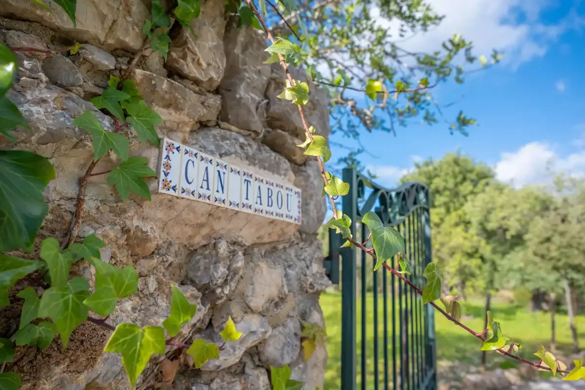 Un muro de piedra con una placa con el nombre de la propiedad, junto a una verja metálica y vegetación.
