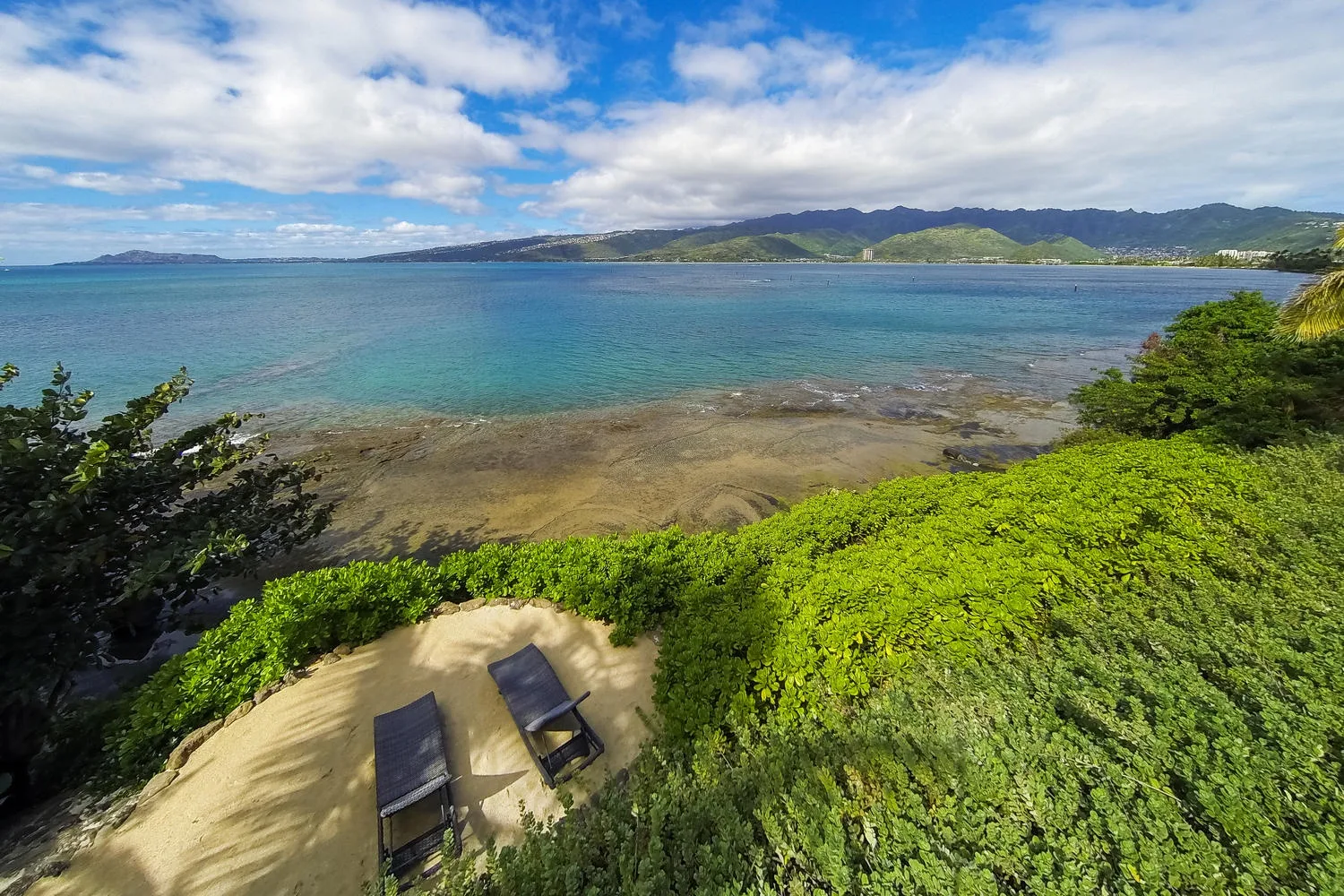 A coastal bay stretches beyond green vegetation, with two lounge chairs on a sandy patch.