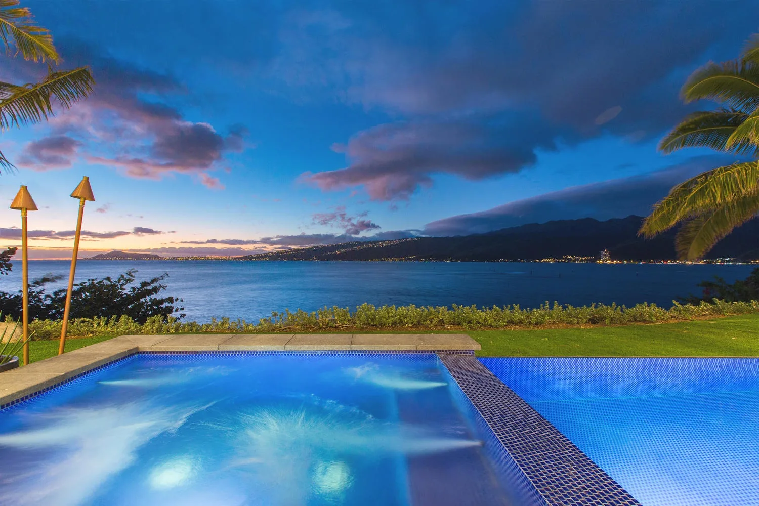 An outdoor swimming pool overlooks a calm bay beneath a colorful evening sky.