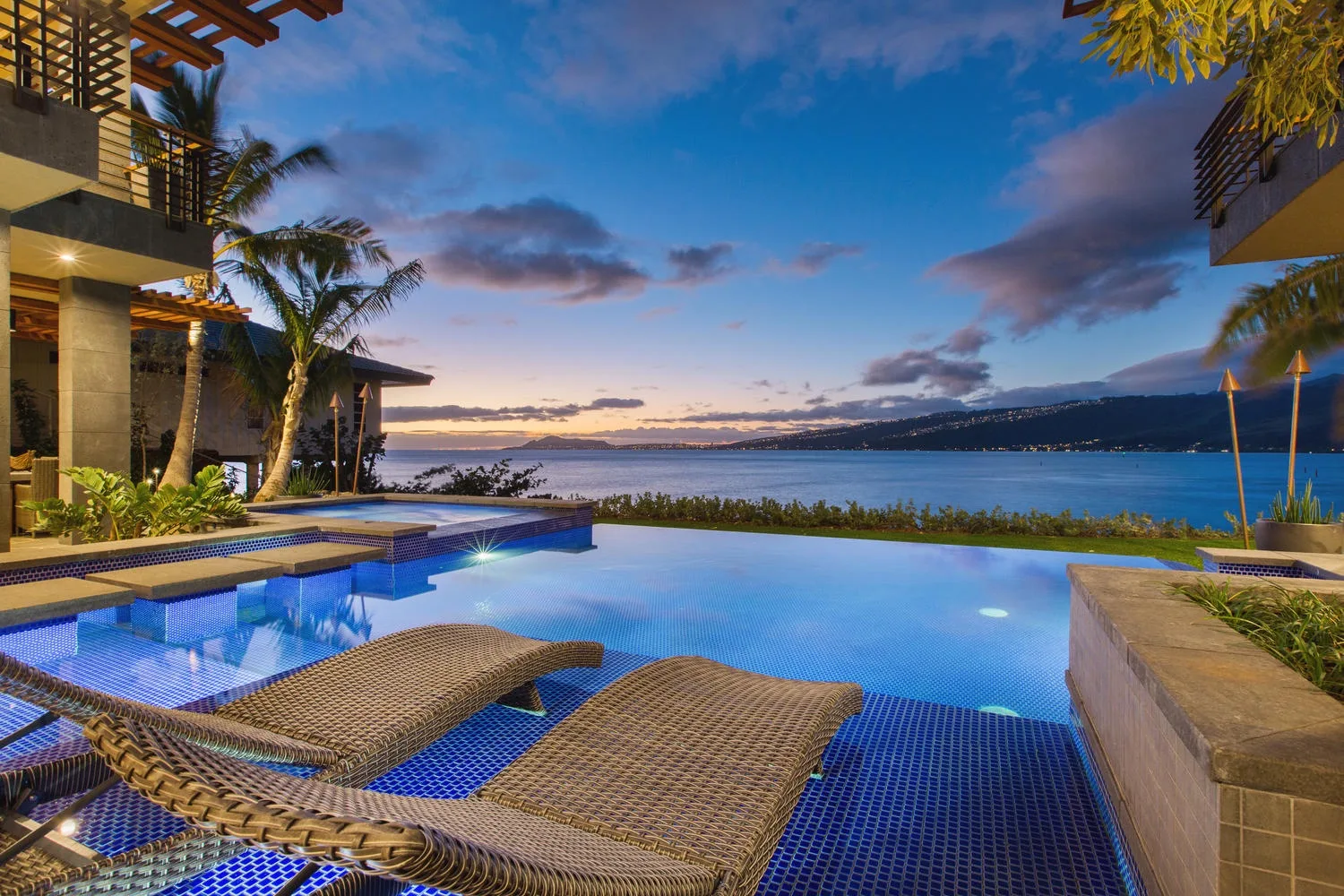 An outdoor swimming pool overlooks a bay at sunset, with loungers beside the water.